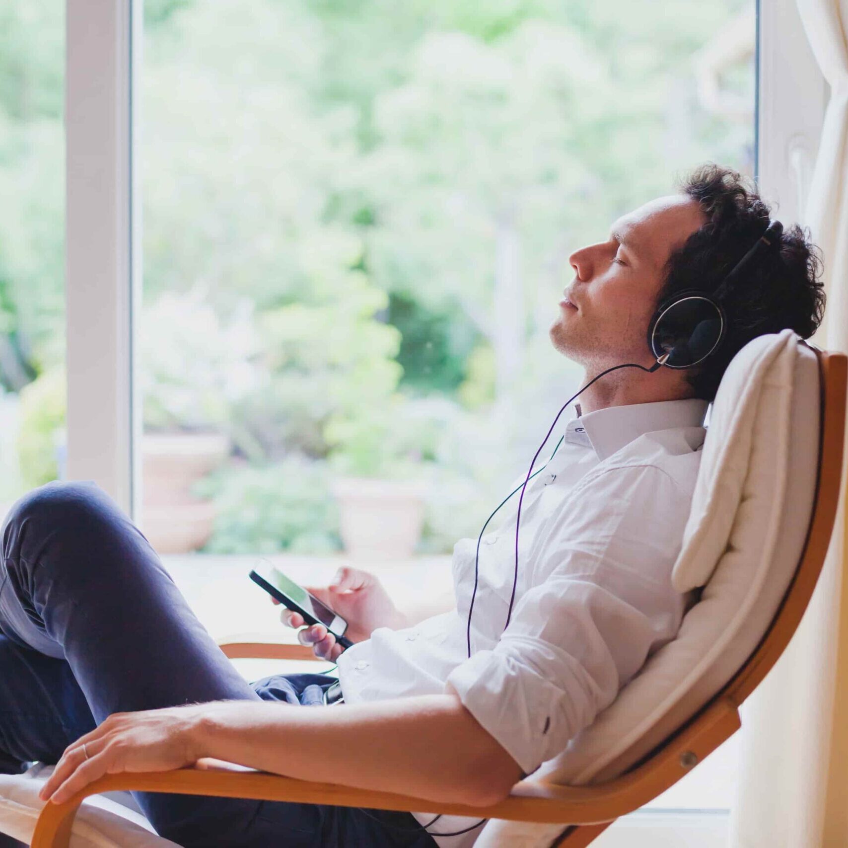 Man in een stoel met koptelefoon, die muziek luistert en door een groot raam naar het groene landschap kijkt.
