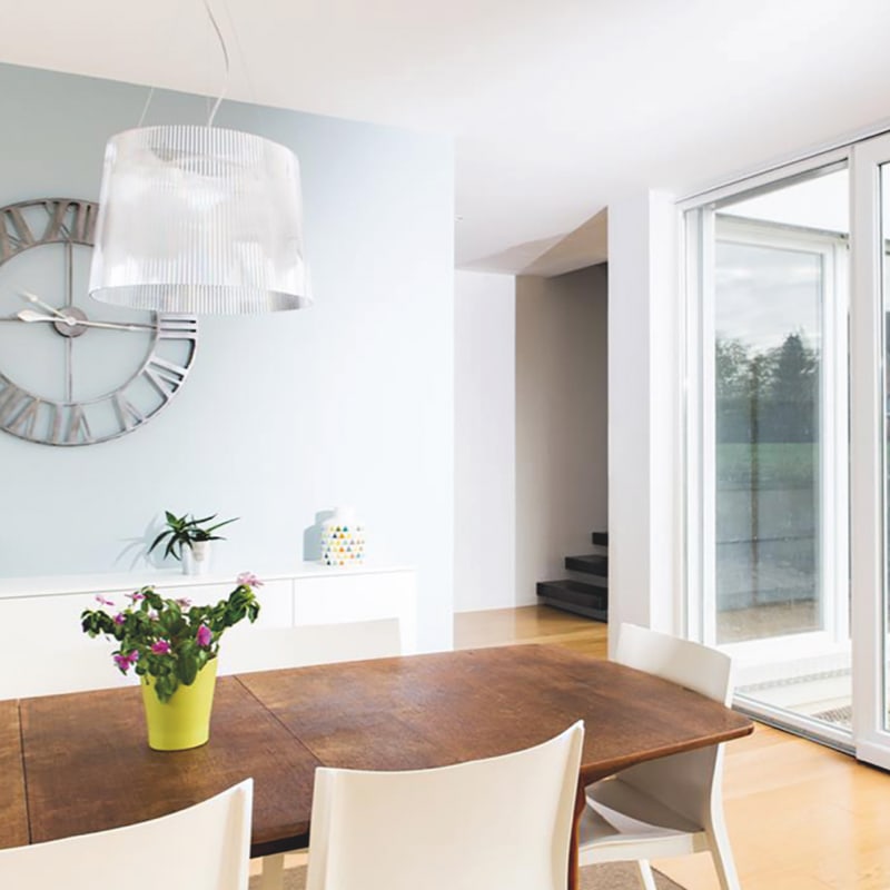 Een moderne eetkamer met een grote houten tafel, witte stoelen, een wandklok en een schuifdeur met uitzicht naar buiten.